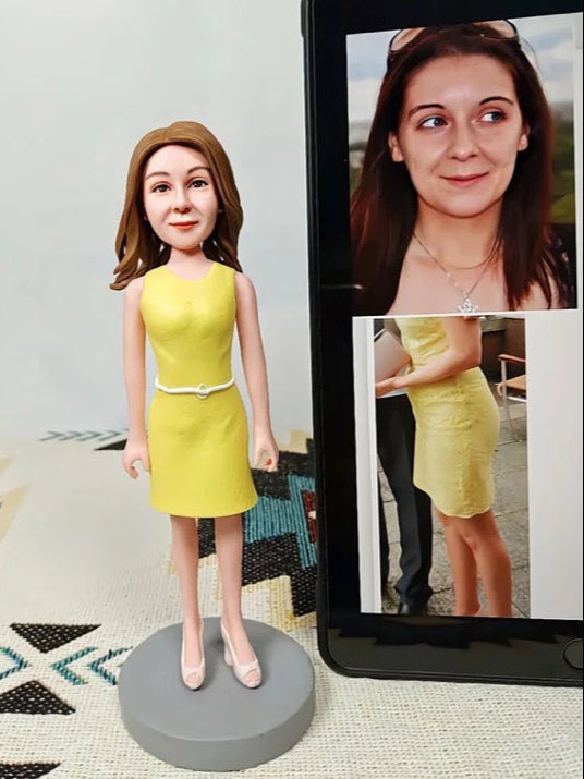 Custom bobblehead of a woman in a yellow dress next to a tablet displaying a photo of the same woman.