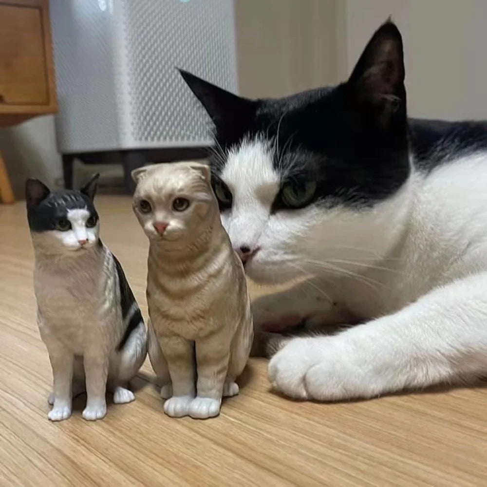 Real cat sitting next to two cat figurines on a wooden floor.